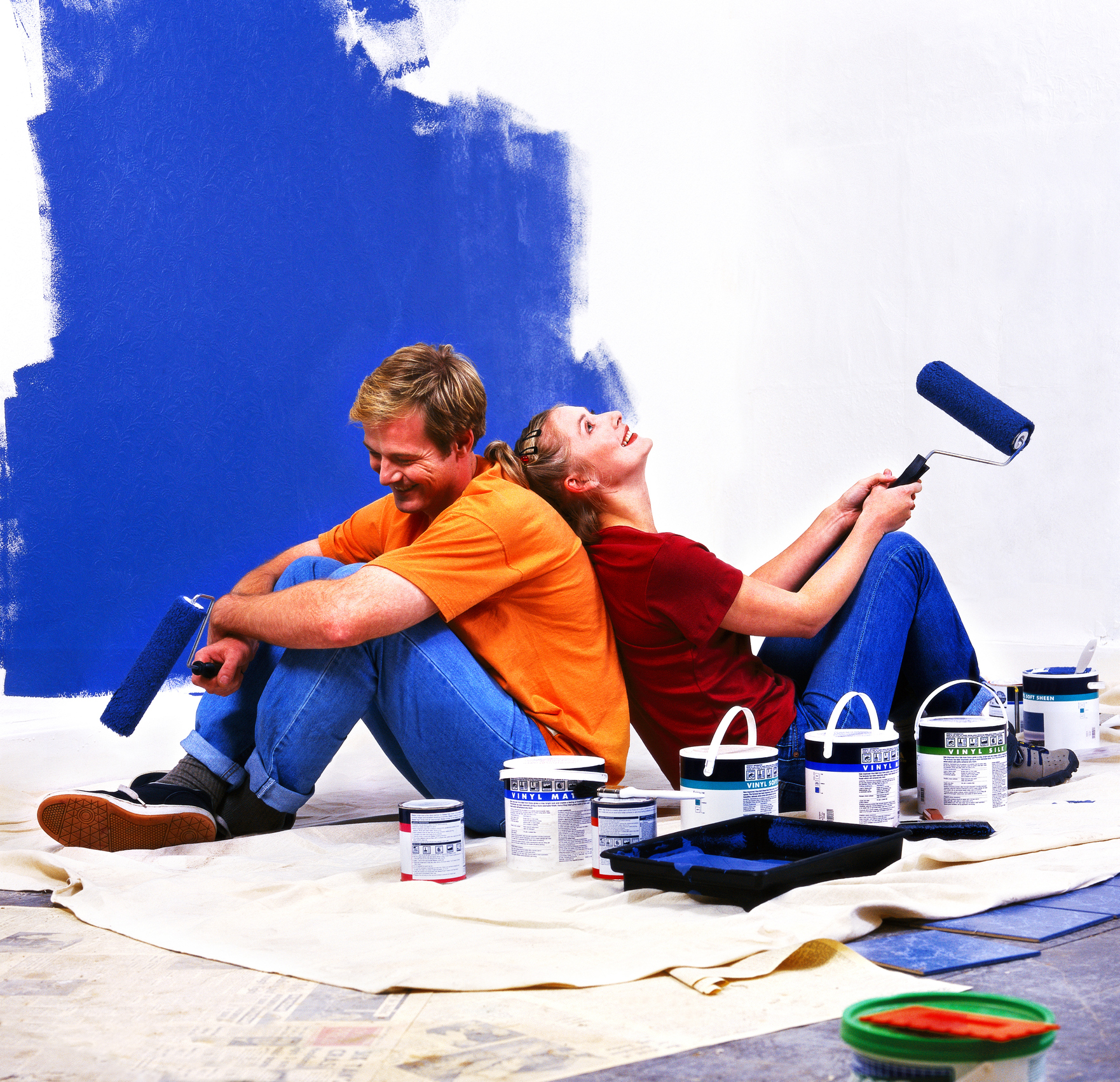 a couple sitting back to back with roller brushes of paint in hand